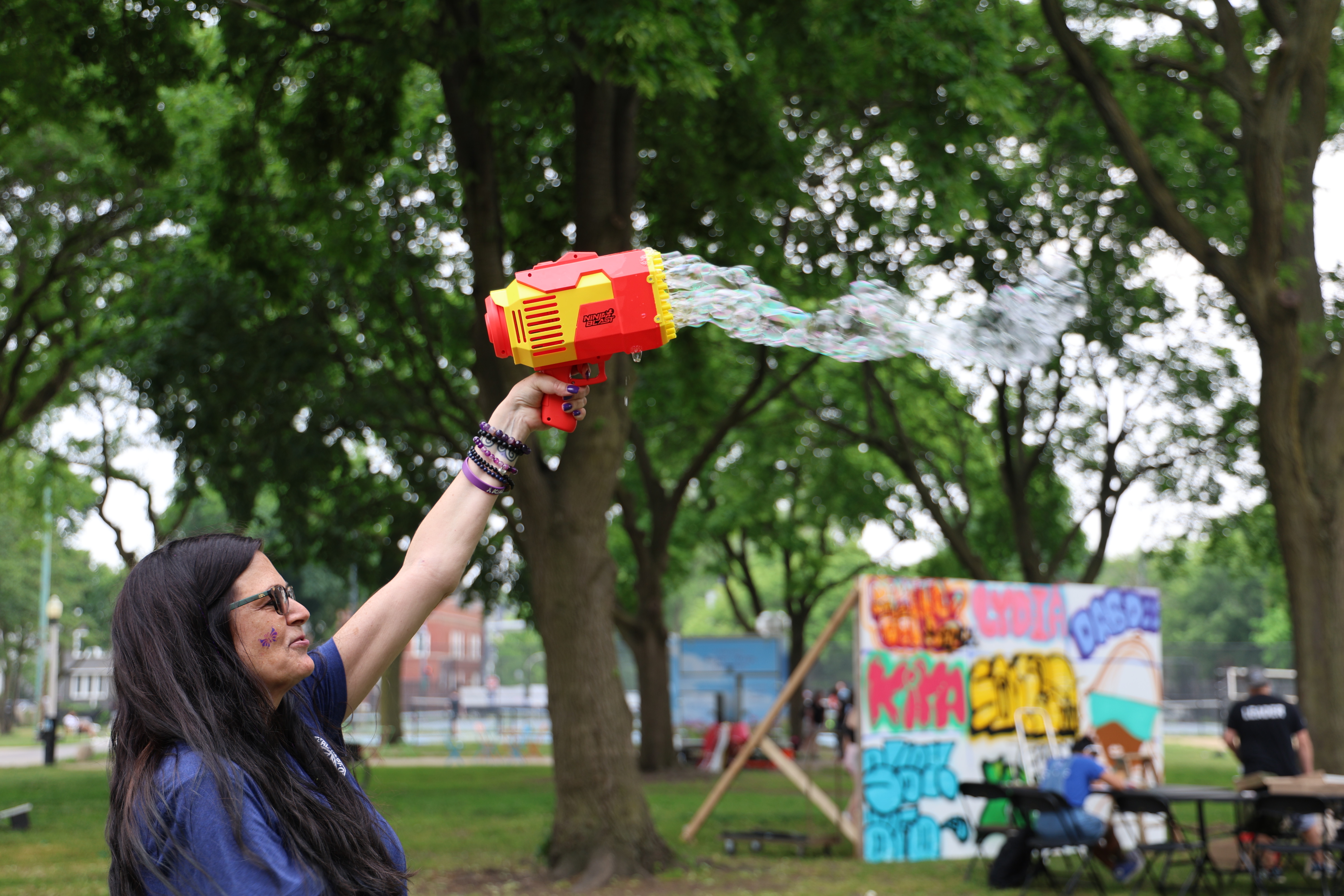 Woman aims a red and yellow bubble gun, sending a stream of bubbles into the air at a park.